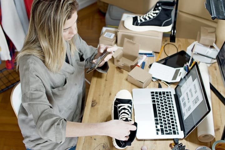 Woman photographing shoes for online sale in home workspace with laptop and packages.